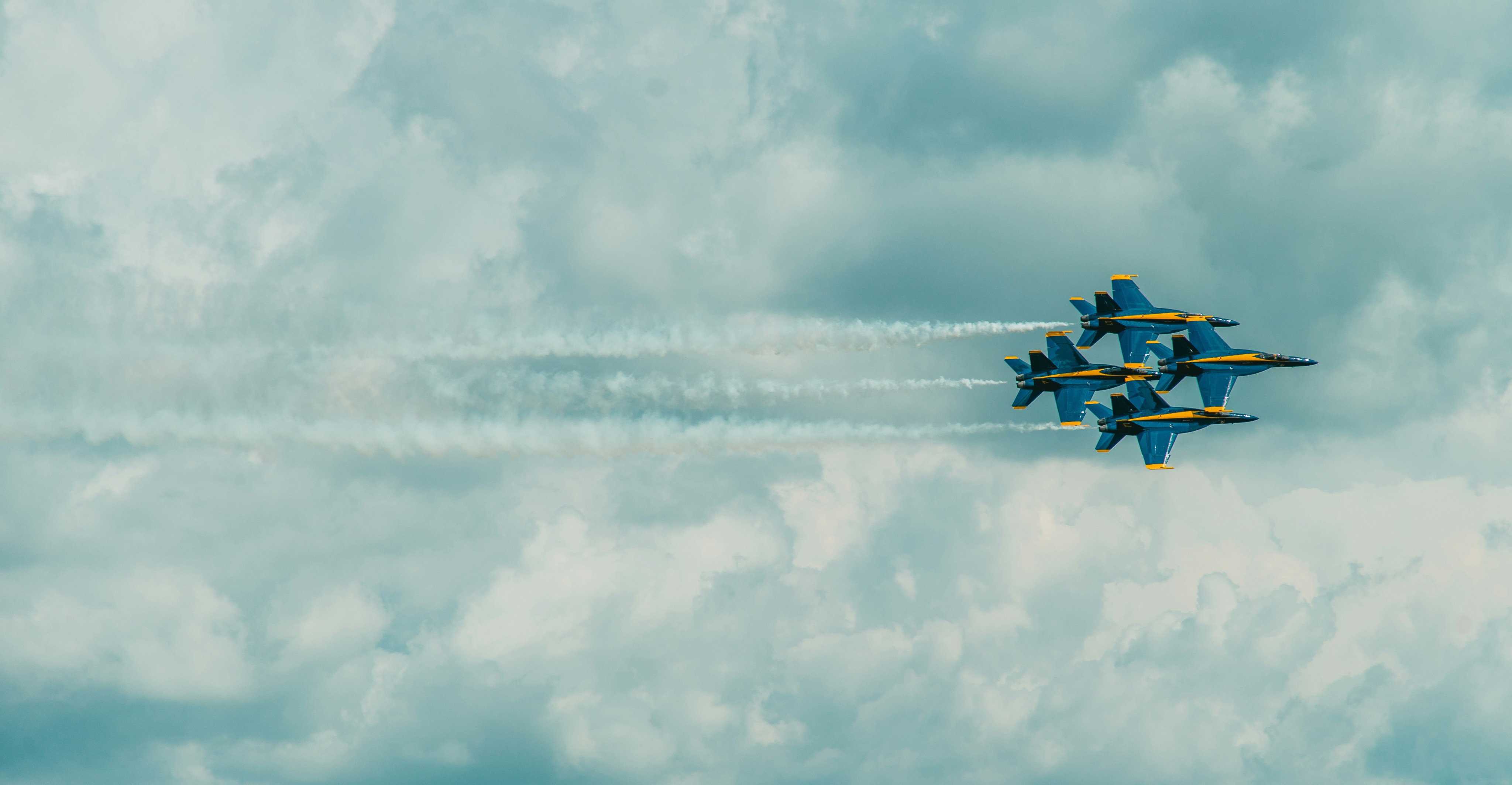 Four F/A-18 Hornets of the US Navy Blue Angels in tight diamond formation against a high overcast sky, contrails trailing.