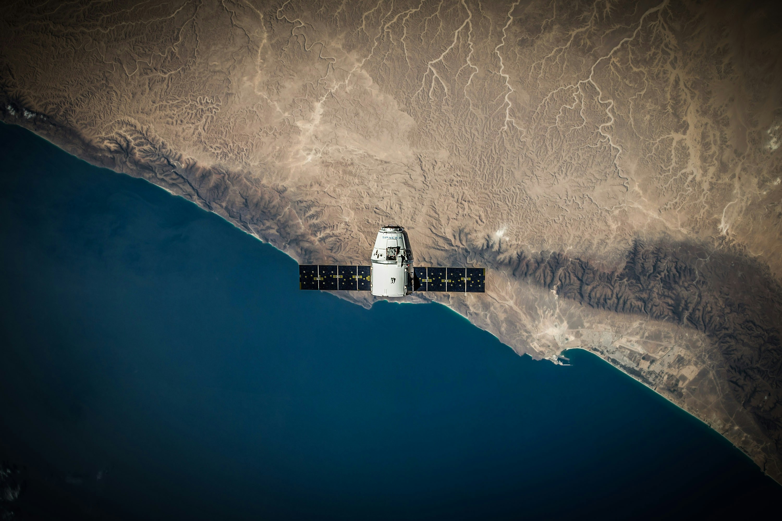SpaceX Dragon capsule with solar arrays deployed, photographed from low Earth orbit looking down at the Baja California coastline.